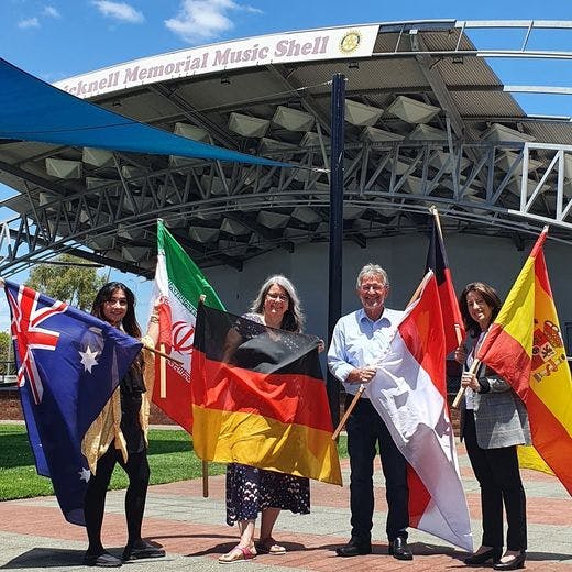 Four people holding the Australian and various international flags in front of the Graham Bricknell Memorial Music Shell.