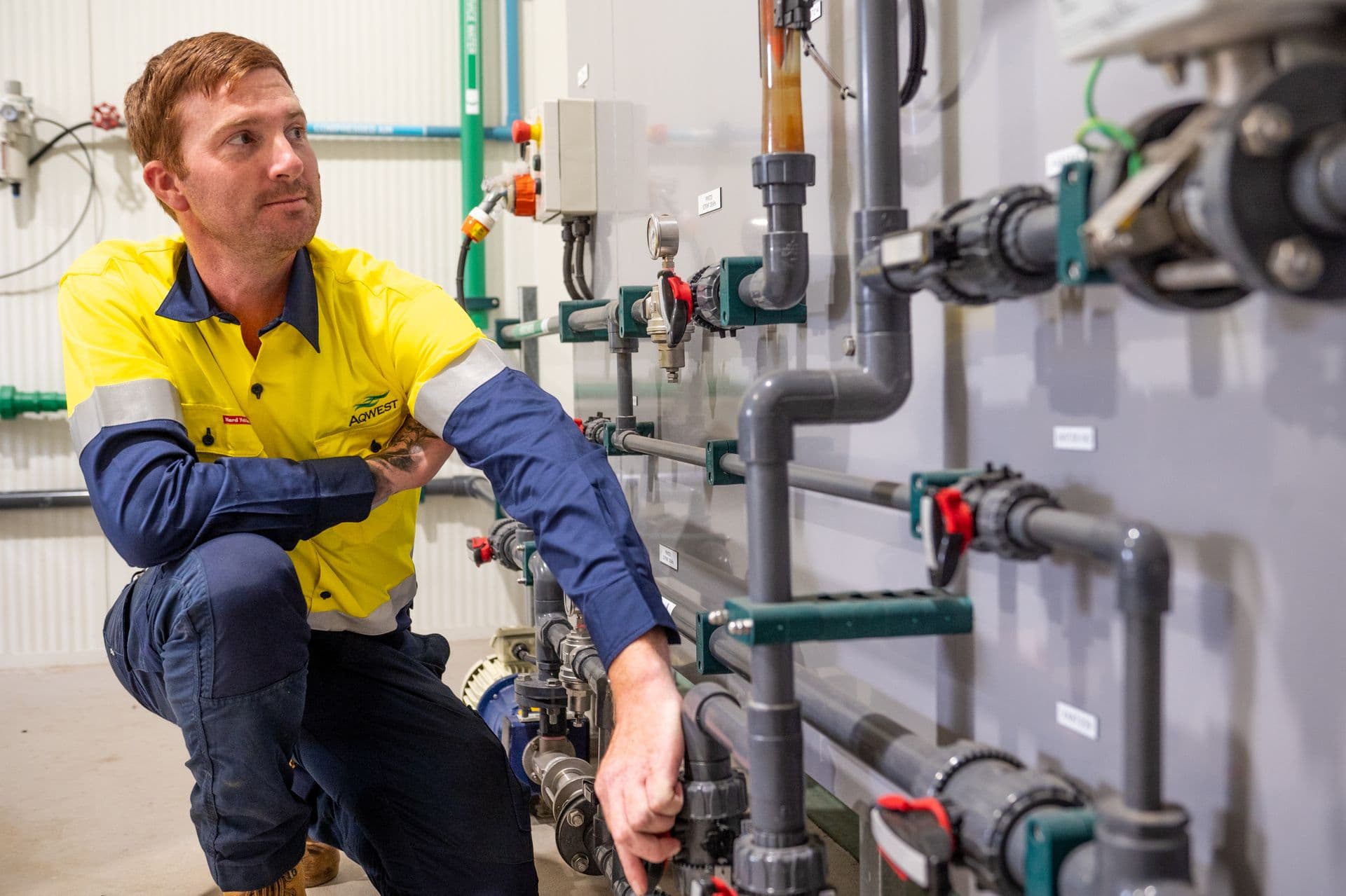 man kneeling on one knee next to a wall covered in pipes and valves.