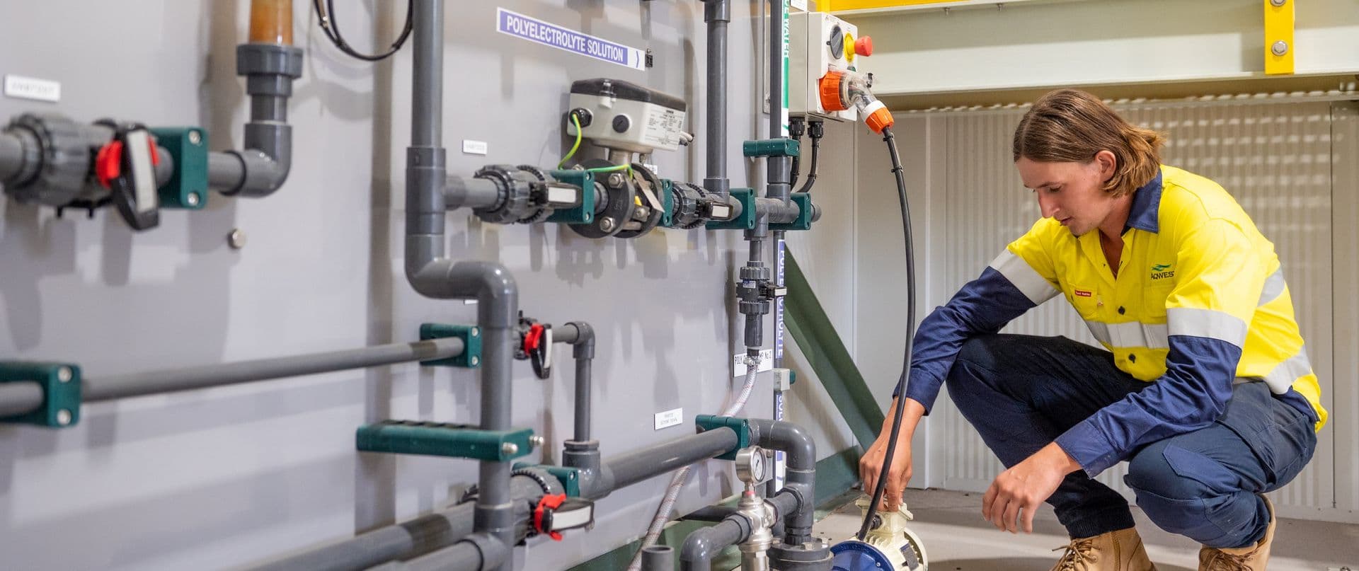 Person crouching in front of pump, a wall of pipe fittings in foreground.