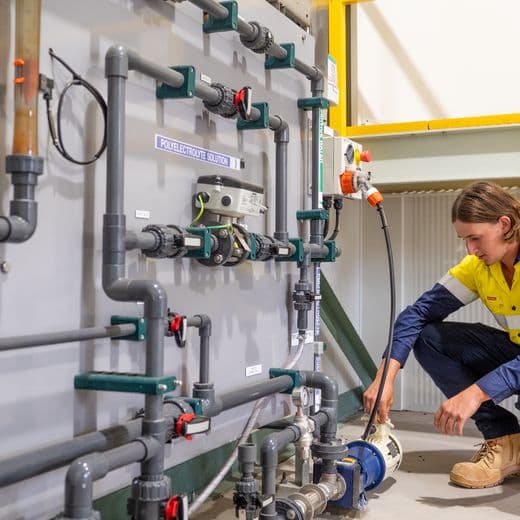 Person crouching in front of pump, a wall of pipe fittings in foreground.