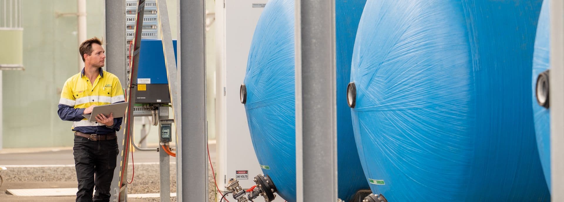 Person in high vis with laptop walking beside blue water tanks.