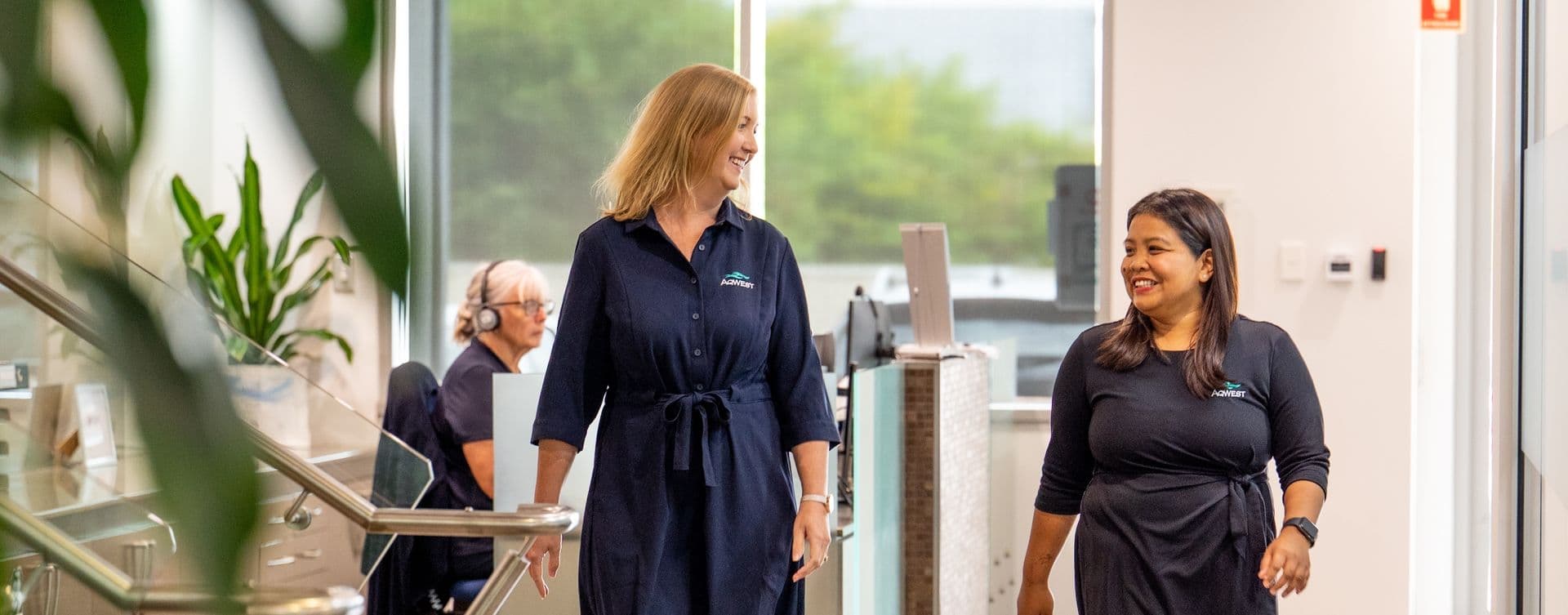 Two people walking side by side in an office, talking and smiling, reception in the background.