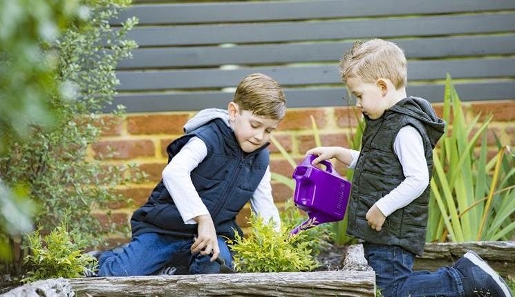 Two children with trowel and a watering can sitting next to a plant.